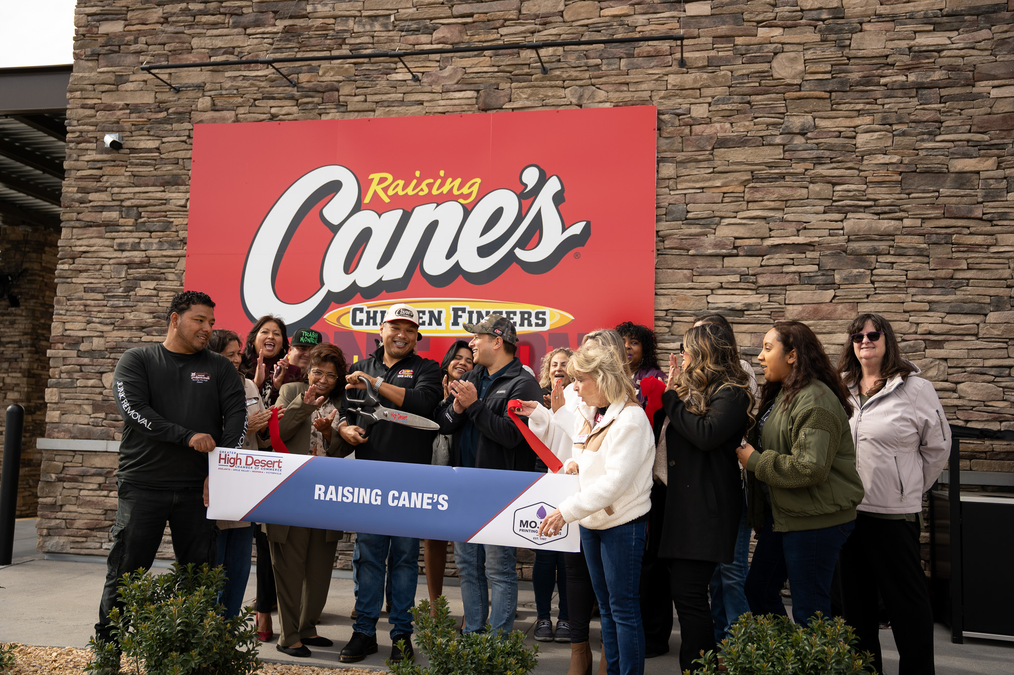 Victorville mayor and community leaders participate in a ribbon-cutting ceremony at the Raising Cane’s grand opening, celebrating the official opening with staff and attendees gathered outside the restaurant.