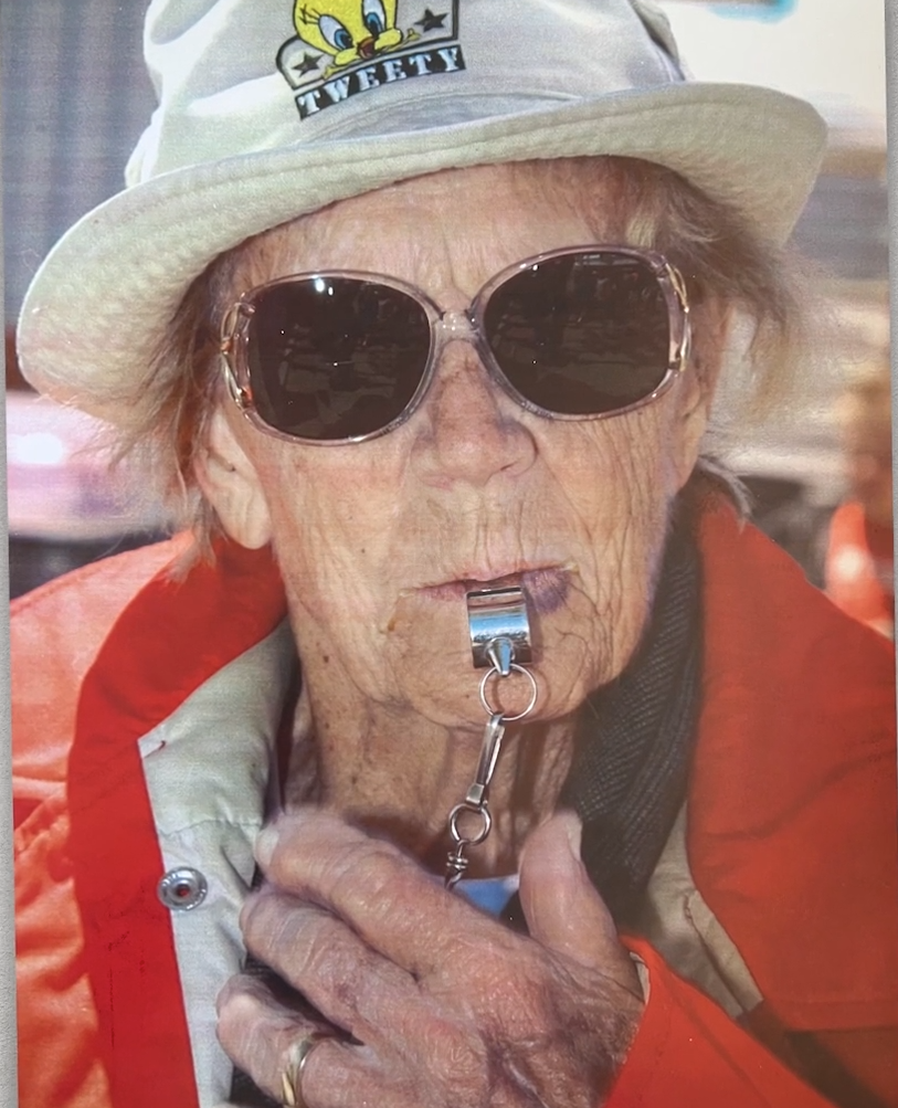 Woman in a red jacket and sunglasses blowing a whistle.