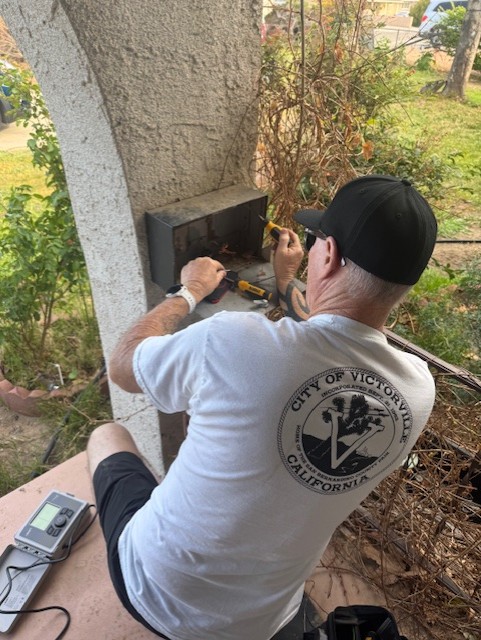 Man wearing a cap and white shirt using tools to repair or adjust an outdoor irrigation control box on a stucco wall.