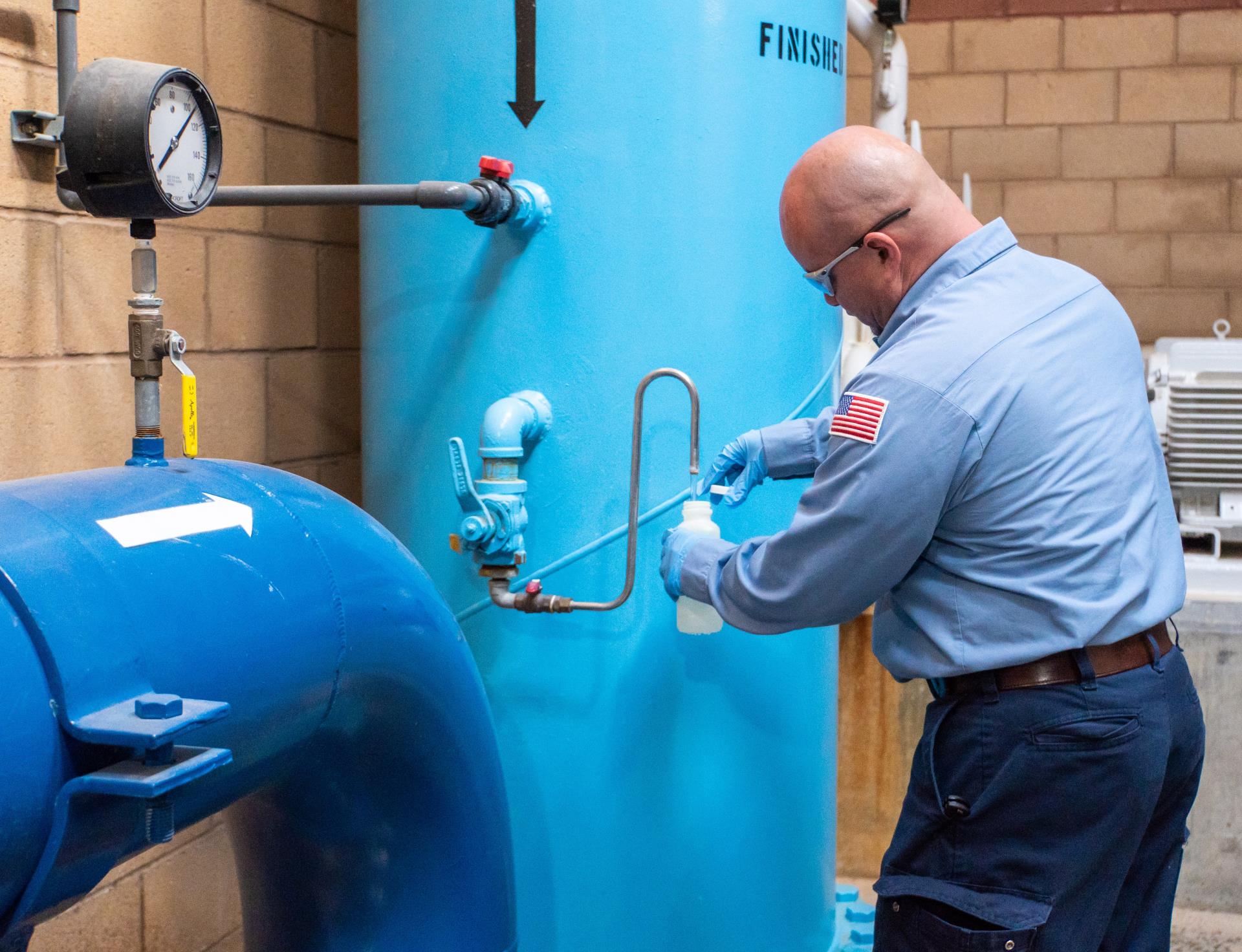 Worker in uniform with American flag patch uses a small container to take a liquid sample from piping connected to a large blue tank with pressure gauges