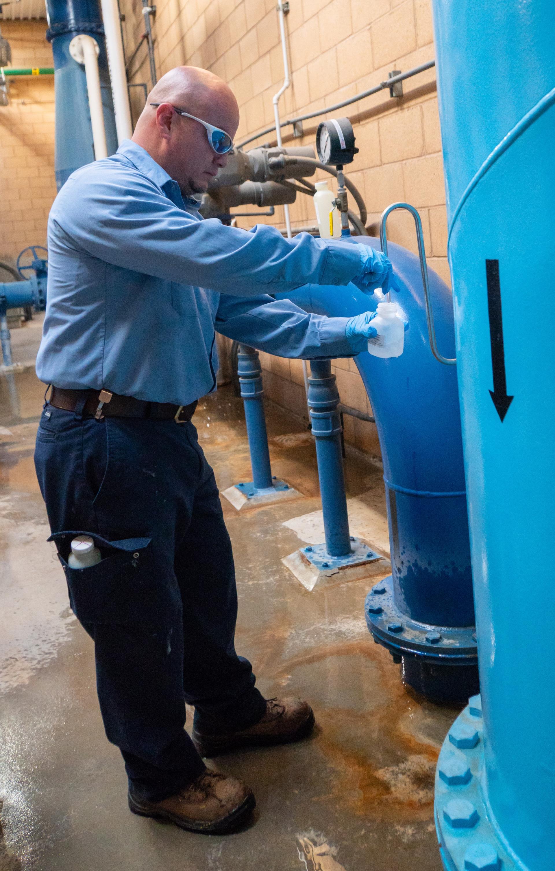 Technician wearing gloves and safety glasses collects a water sample from a valve on a large blue industrial tank in a utility room