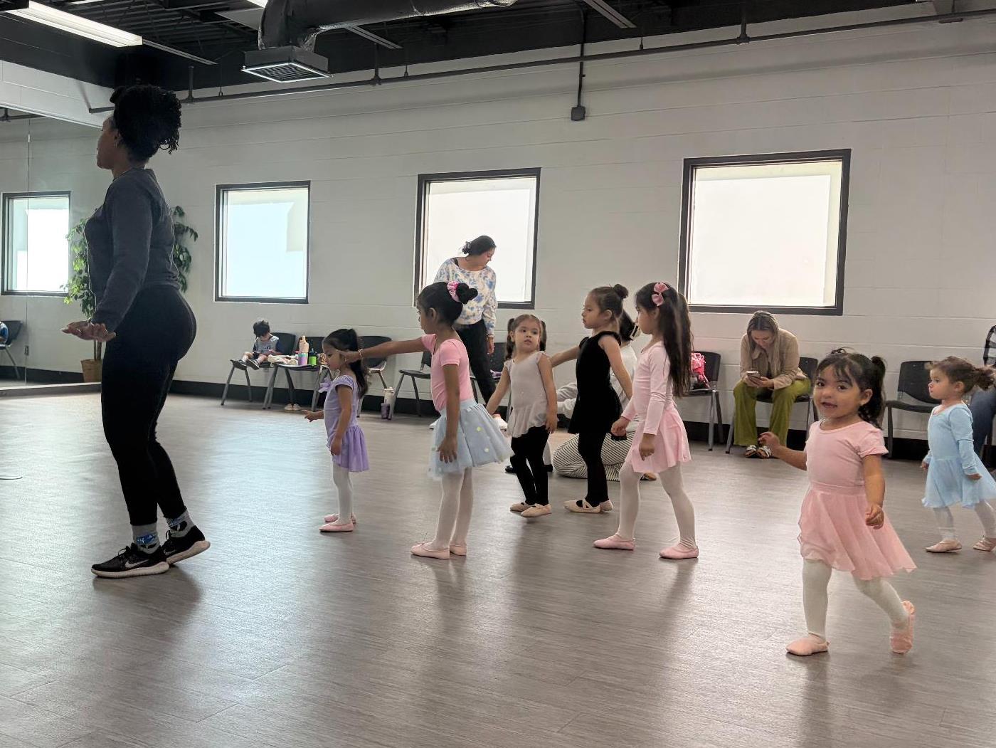 A dance room with an instructor next to a group of young children practicing a dance in front of a mirror.