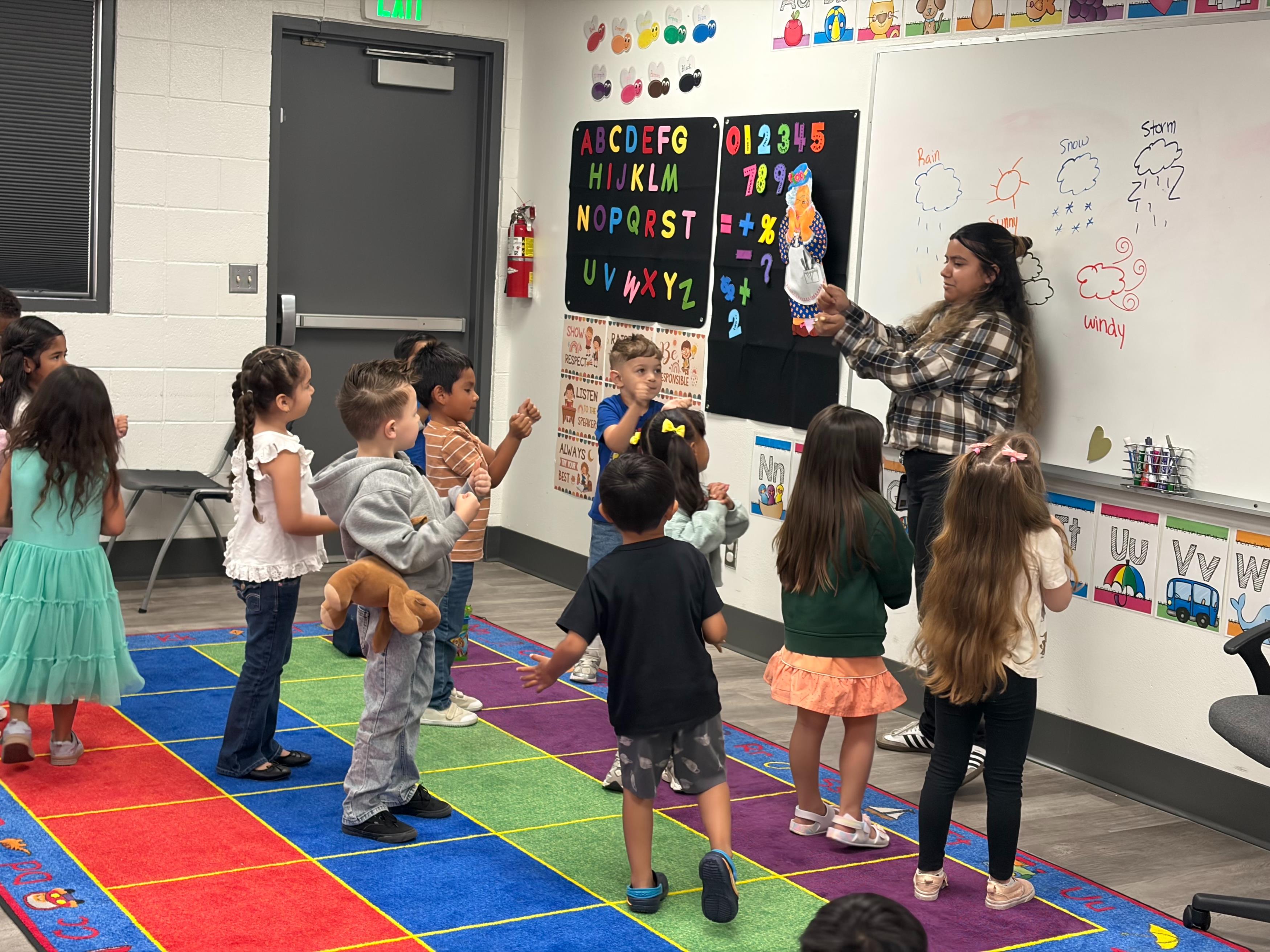 A group of preschool students learning a dance with their teacher in the classroom.